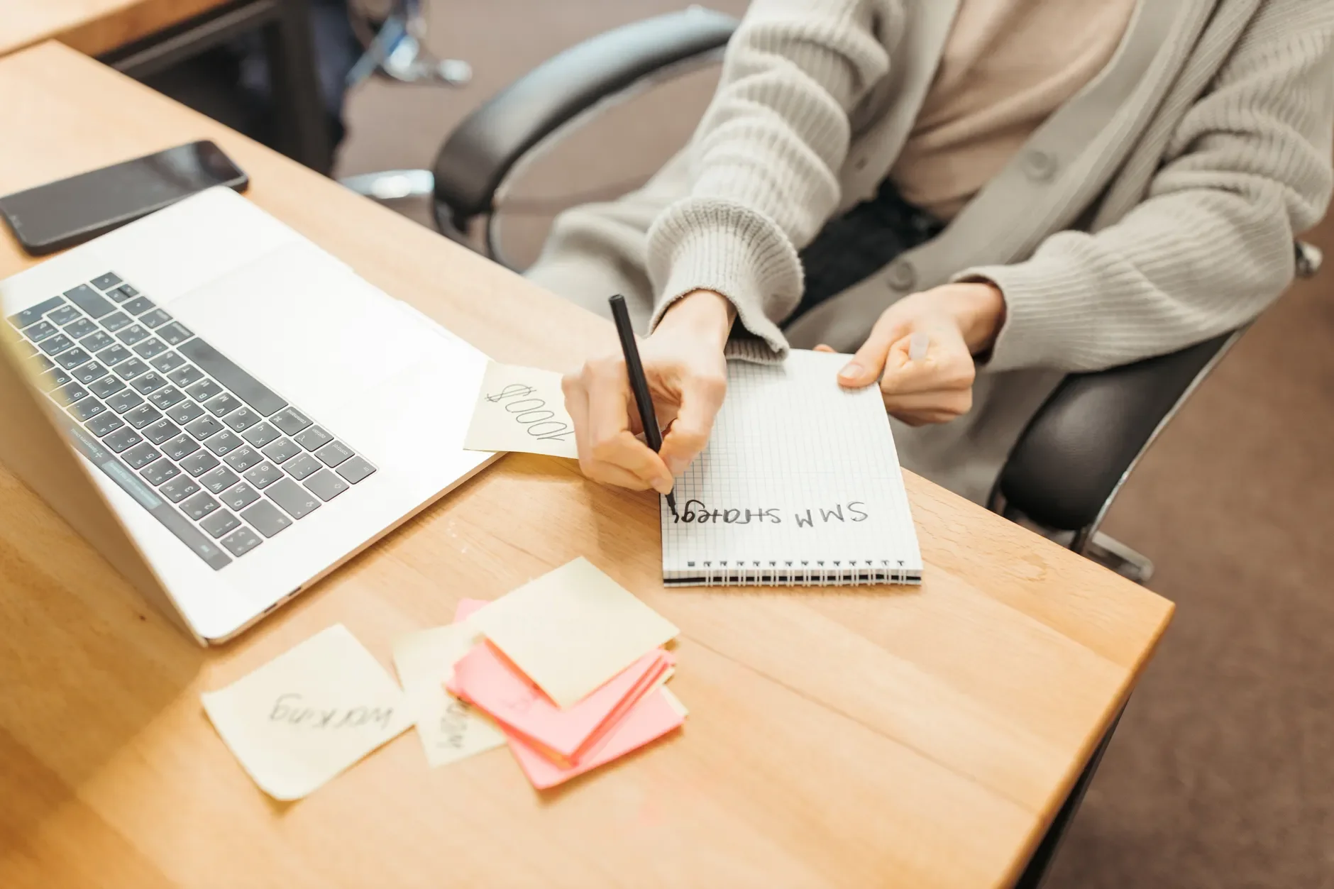 Eine Person schreibt mit einem Stift in einen Spiralblock neben einem geöffneten Laptop auf einem hellen Holzschreibtisch.