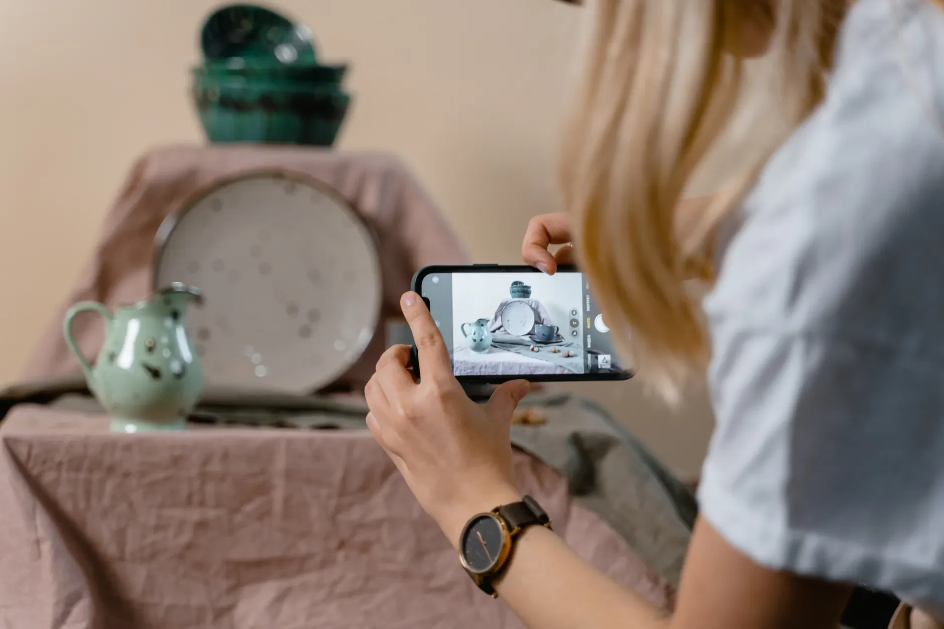 Eine Person fotografiert mit einem quer gehaltenen Smartphone arrangiertes Keramikgeschirr auf einem mit Stoff bedeckten Tisch.