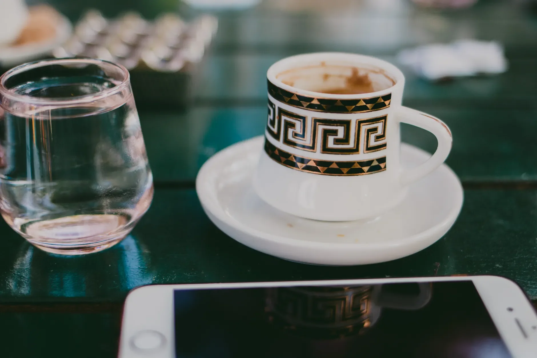 Gemusterte Kaffeetasse auf einer Untertasse neben einem Wasserglas und einem Smartphone auf einem dunkelgrünen Tisch.