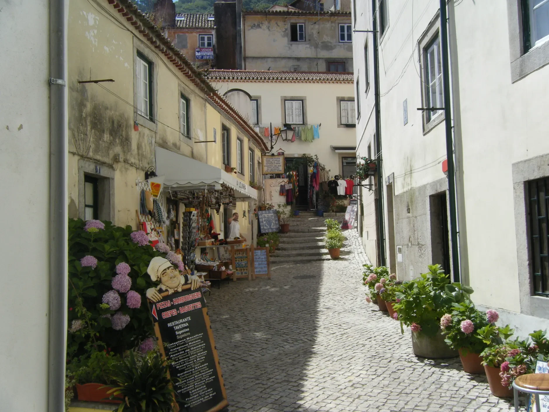 Schmale gepflasterte Gasse mit kleinen Geschäften, hellen Fassaden und blühenden Pflanzen in Töpfen.